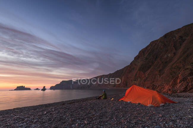 Femme âgée par tente assise sur la plage au coucher du soleil — Photo de stock