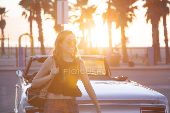 Усміхнена молода жінка за класичною машиною на заході сонця — стокове фото