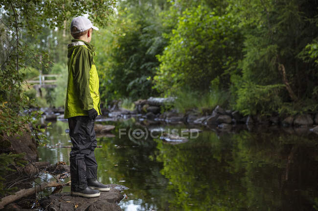 Мальчик в шапке и куртке, стоящий на камне у ручья — стоковое фото