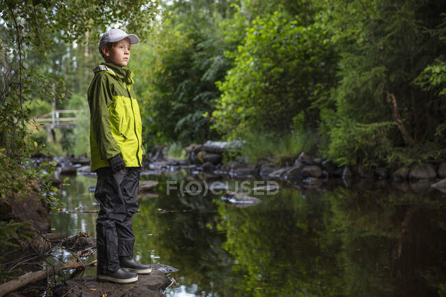 Мальчик в шапке и куртке, стоящий на камне у ручья — стоковое фото