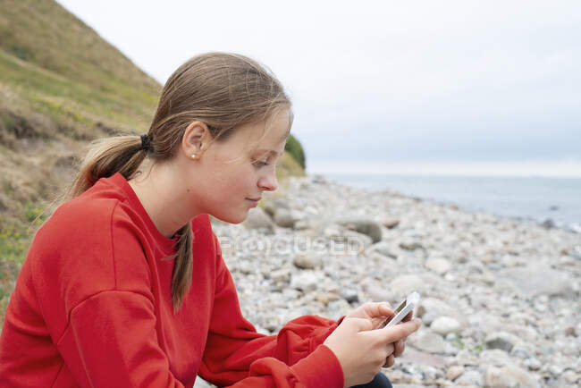 Adolescente chica mensajes de texto en la orilla del río - foto de stock