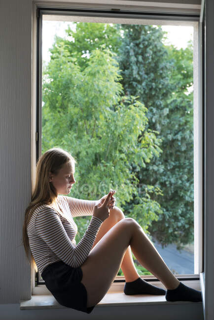 Mensajería de texto de chica adolescente en alféizar ventana - foto de stock