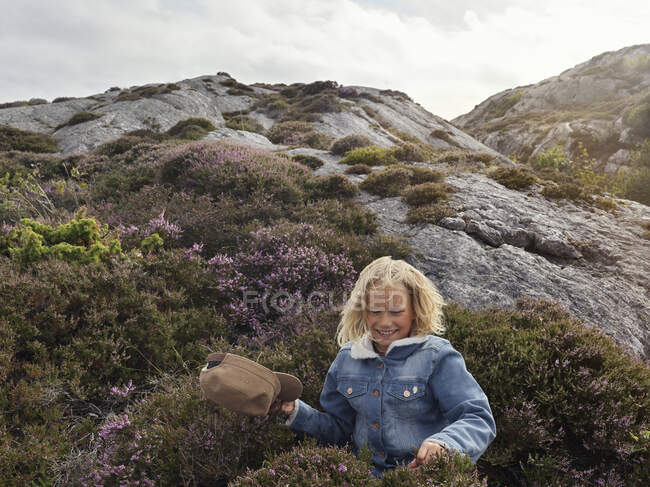 Menina sorridente em jaqueta de ganga em pé em arbustos por rocha — Fotografia de Stock