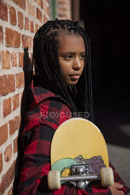 Teenage girl holding skateboard while leaning on brick wall — Stock Photo
