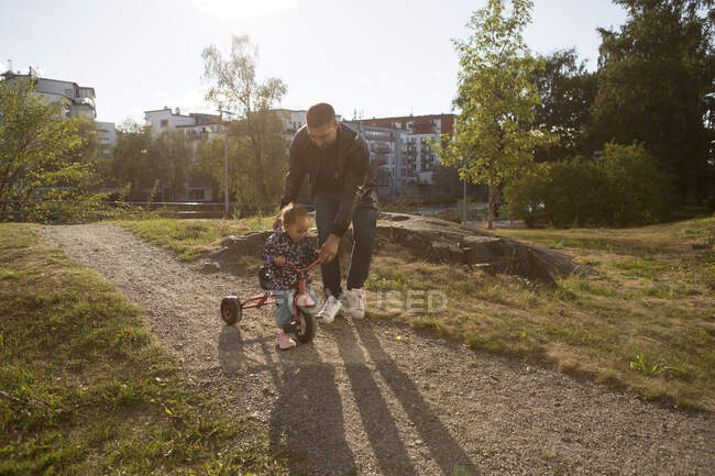 Человек помогает своей дочери ездить на трехколесном велосипеде в парке — стоковое фото
