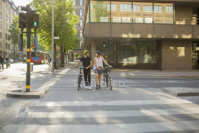 Joven hombre y mujer caminando bicicletas en el cruce peatonal - foto de stock