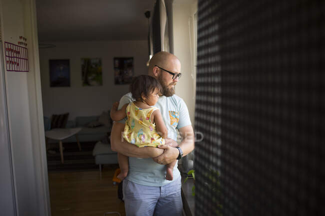 Мужчина держит дочь, глядя в окно — стоковое фото