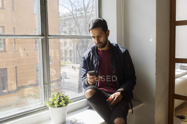 Jeune homme utilisant un smartphone sur le rebord de la fenêtre — Photo de stock