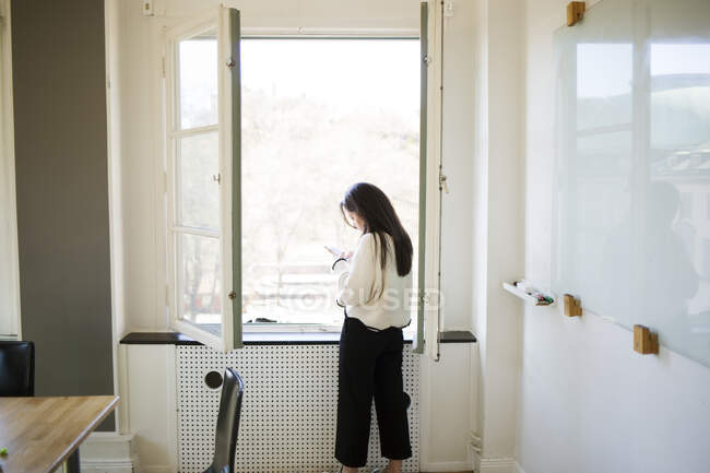 Jovem mulher usando smartphone por janela — Fotografia de Stock