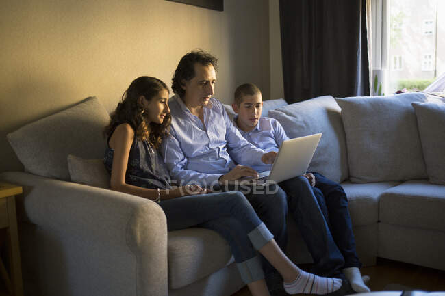 Hombre con portátil sentado con sus hijos en el sofá - foto de stock