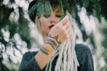 Young woman with dreadlocks and henna tattoos touching coniferous tree branch — Stock Photo