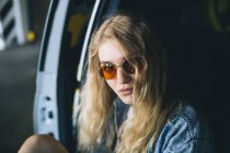 Блондинка в солнечных очках сидит у машины и смотрит в камеру — стоковое фото