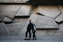 Повнометражний знімок двох жінок у класичних костюмах, позує на геометричну бетонну стіну на відкритому повітрі — стокове фото