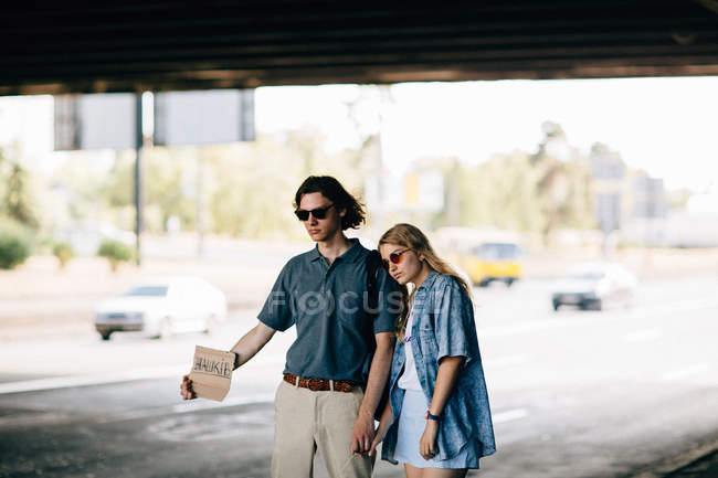 Young couple in sunglasses hitchhiking with cardboard sign — Stock Photo