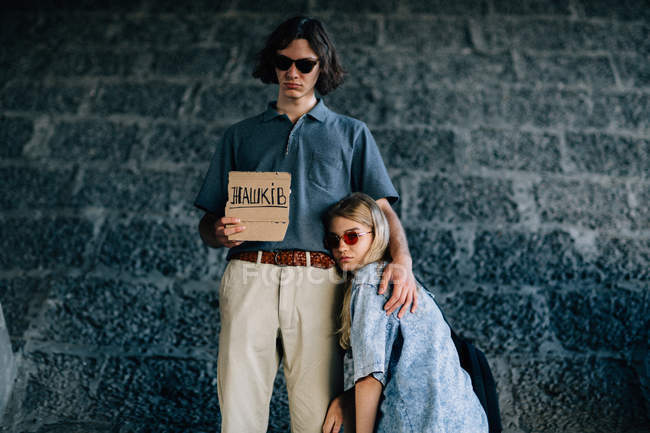 Stylish couple in sunglasses posing with hitchhiking cardboard plate — Stock Photo