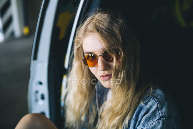 Blonde girl in sunglasses sitting by car and looking at camera — Stock Photo