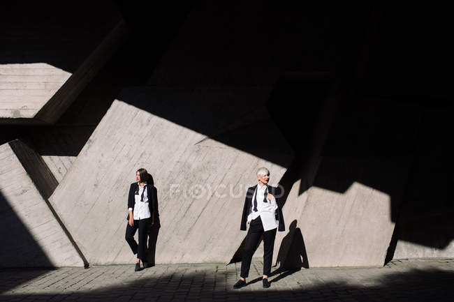 Tiro de longitud completa de dos mujeres en trajes clásicos posando contra pared de hormigón geométrico al aire libre - foto de stock