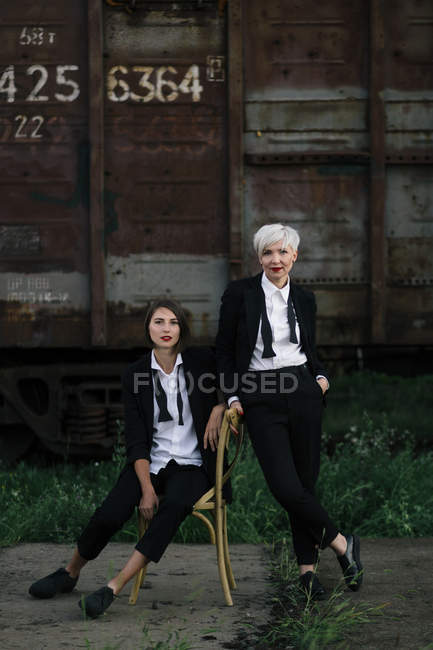 Tiro de longitud completa de dos mujeres posando en la estación de tren, una sentada en la silla y la otra de pie cerca - foto de stock