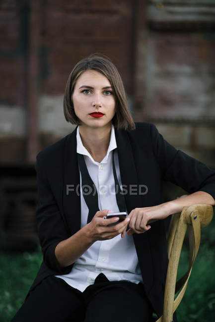 Portrait de jeune femme utilisant un smartphone et regardant la caméra à la gare en arrière-plan — Photo de stock