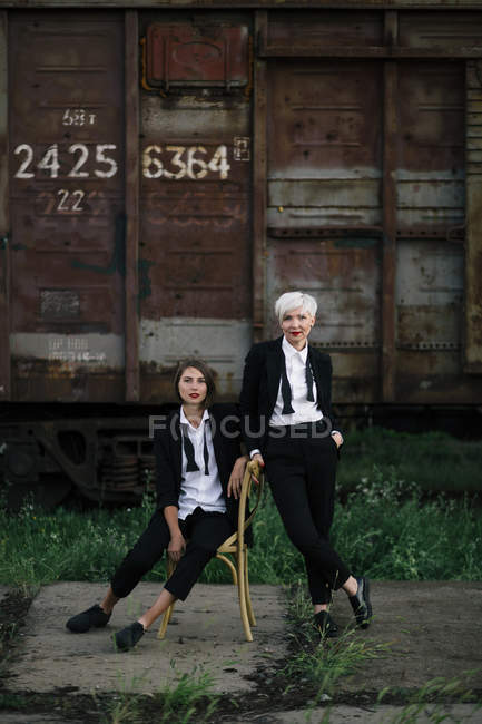 Tiro de longitud completa de dos mujeres posando en la estación de tren, una sentada en la silla y la otra de pie cerca - foto de stock