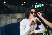 Молодий чоловік п'є пиво в барі з жіночою рукою, регулюючи сонцезахисні окуляри — стокове фото