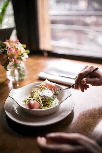 Vue recadrée de la personne tenant fourchette par des crêpes de pommes de terre avec jambon, sauce et herbes dans un bol — Photo de stock