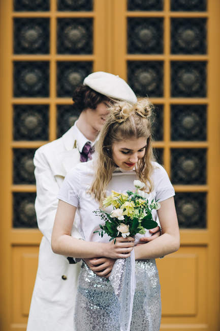 Молодая обнимающая пара позирует с свадебным букетом против двери здания — стоковое фото