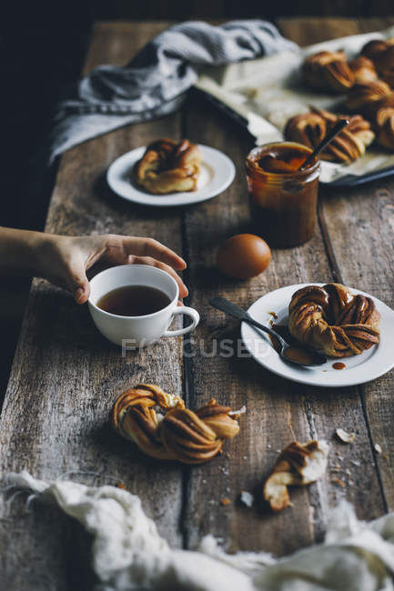 Main féminine avec tasse de thé et petits pains à la cannelle suédoise sur table en bois rustique — Photo de stock