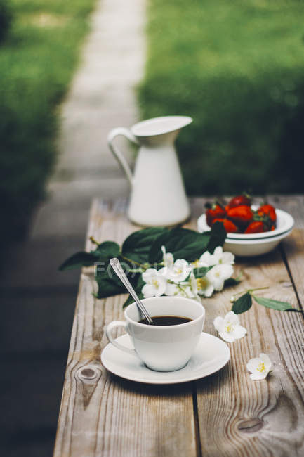 Чашка кофе на деревянном столе в саду со свежими цветами — стоковое фото