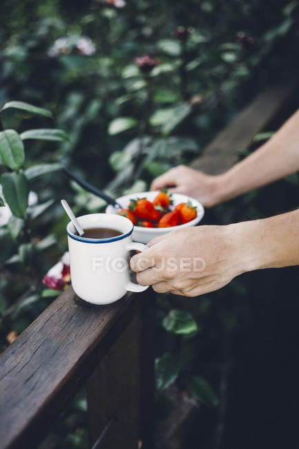 Human hands holding cup of black tea and bowl of fresh strawberries on wooden plank outdoors — Stock Photo