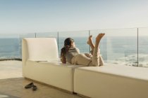 Жінка на розкішному балконі, що розслабляється, лежачи на лавці, дивлячись на сонячний вид на океан — стокове фото