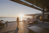Femme marchant sur le coucher du soleil moderne, maison de luxe vitrine patio extérieur avec vue sur l'océan — Photo de stock