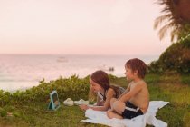 Мальчик и девочка брат и сестра смотрят видео на цифровой планшет в траве с видом на океан — стоковое фото