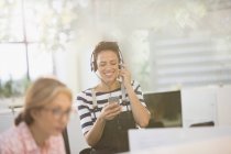 Femme d'affaires créative souriante écoutant de la musique avec écouteurs et téléphone intelligent — Photo de stock