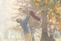 Pai levantando filho sobrecarga em madeiras com folhas de outono — Fotografia de Stock