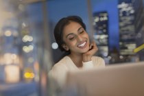 Mujer de negocios sonriente trabajando hasta tarde en el ordenador portátil en la oficina por la noche - foto de stock