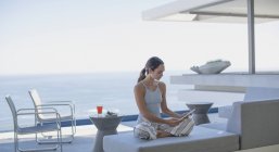 Mujer en pijama usando tableta digital en moderno, casa de lujo escaparate patio exterior con vista al mar - foto de stock