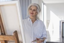 Smiling mature woman with palette painting at canvas on easel — Stock Photo