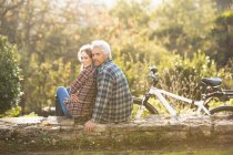 Portrait affectionate couple with bicycle resting on stone wall in autumn park — Stock Photo