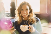 Ritratto donna sorridente con auricolari che ascolta musica su lettore mp3 e beve cappuccino nel caffè — Foto stock