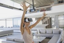 Femme énergique utilisant des lunettes de simulateur de réalité virtuelle avec les bras levés dans le salon moderne et luxueux de vitrine de la maison — Photo de stock