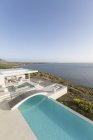 Sunny, tranquillo moderno lusso casa vetrina piscina a sfioro con passerella e vista sull'oceano sotto il cielo blu — Foto stock