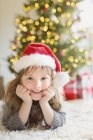 Портрет усміхненої дівчини в капелюсі Санта на килимі у вітальні з ялинкою — стокове фото