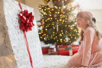 Girl smiling in anticipation at large Christmas gift near Christmas tree — Stock Photo