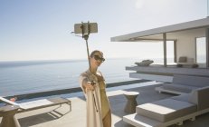 Woman in bathing suit taking selfie with selfie stick on modern, luxury home showcase exterior deck with ocean view — Stock Photo