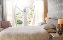 Donna in accappatoio aprendo tende camera da letto — Foto stock