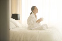 Donna sorridente in accappatoio bere caffè a gambe incrociate sul letto — Foto stock