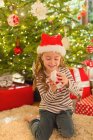 Smiling girl in Santa hat opening gift in front of Christmas tree — Stock Photo