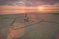 Garçon et fille frère et soeur dessinant en forme de coeur dans le sable sur la plage tranquille coucher de soleil — Photo de stock