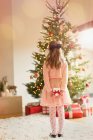 Chica en vestido rosa sosteniendo regalo de Navidad delante del árbol de Navidad - foto de stock